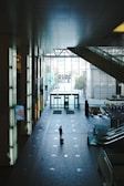 A lone individual stands in a spacious, modern indoor space, pulling a suitcase. The area is characterized by high ceilings, large glass windows allowing natural light to enter, and a tiled floor with a grid-like pattern. Escalators are visible on the right, and a minimalist electronic display screen on the left. The atmosphere is quiet and somewhat solitary.