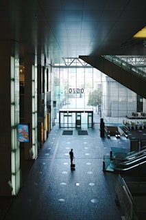 A lone individual stands in a spacious, modern indoor space, pulling a suitcase. The area is characterized by high ceilings, large glass windows allowing natural light to enter, and a tiled floor with a grid-like pattern. Escalators are visible on the right, and a minimalist electronic display screen on the left. The atmosphere is quiet and somewhat solitary.