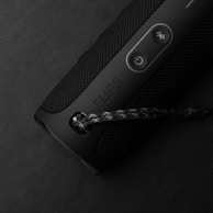 A black portable Bluetooth speaker with a textured surface and control buttons. It features a power button and a Bluetooth pairing button. The speaker has a carrying strap attached to it.
