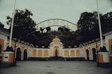 An entrance gate with the word 'SKANSEN' displayed prominently above. The architecture is a blend of arches and classical details with mustard yellow and white colors. Flanking the entrance are orange-toned walls adorned with classic street lamps. Large trees tower behind this historical entrance.