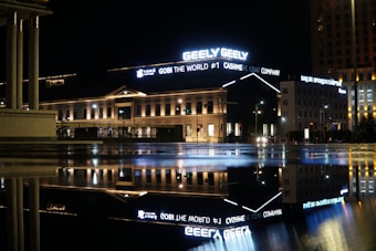 A nighttime cityscape featuring an illuminated building with large neon signs, including ads for Geely and a cashmere coat company. The building's elegant architecture is highlighted by warm lighting, and it is reflected in a puddle on the pavement, providing a symmetrical visual effect.