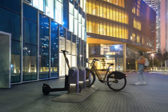 A bicycle and electric scooter are parked at a docking station in a modern urban setting. The scene is set against a backdrop of glass buildings illuminated with various colors, primarily blue and yellow. A person in motion is blurred, indicating movement, and the image is captured in a nighttime environment.