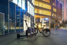 A bicycle and electric scooter are parked at a docking station in a modern urban setting. The scene is set against a backdrop of glass buildings illuminated with various colors, primarily blue and yellow. A person in motion is blurred, indicating movement, and the image is captured in a nighttime environment.