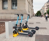 Three electric scooters with blue handles are parked in a designated charging station along a wide, paved sidewalk beside a stone building. Several windows with metal bars are visible on the building. In the background, a cyclist rides down the street lined with trees and other buildings.