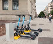Three electric scooters with blue handles are parked in a designated charging station along a wide, paved sidewalk beside a stone building. Several windows with metal bars are visible on the building. In the background, a cyclist rides down the street lined with trees and other buildings.