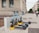 Three electric scooters with blue handles are parked in a designated charging station along a wide, paved sidewalk beside a stone building. Several windows with metal bars are visible on the building. In the background, a cyclist rides down the street lined with trees and other buildings.