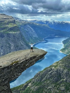 A person stands triumphantly on the edge of a dramatic rock formation overlooking a deep fjord below, surrounded by towering cliffs and expansive mountainous landscape. The sky above is filled with moody clouds, adding a sense of grandeur and adventure to the scene.
