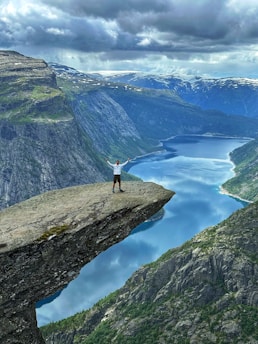 A person stands triumphantly on the edge of a dramatic rock formation overlooking a deep fjord below, surrounded by towering cliffs and expansive mountainous landscape. The sky above is filled with moody clouds, adding a sense of grandeur and adventure to the scene.