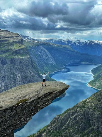 A person stands triumphantly with arms raised on the edge of a prominent cliff overlooking a deep blue fjord surrounded by towering rocky cliffs and snow-capped mountains under a cloudy sky.