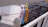 A close-up view of a clothesline with a set of colorful clothespins including pink, blue, and yellow. The background shows a residential building with balconies and windows, suggesting an urban setting. The perspective is from someone standing on a balcony or terrace.