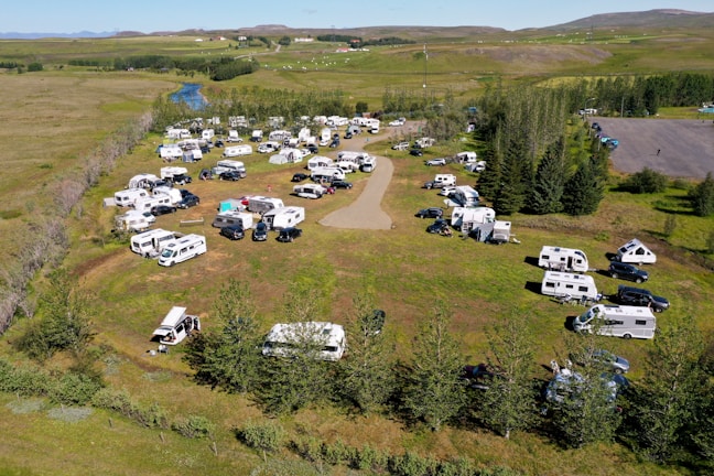 An aerial view of a campsite filled with numerous RVs and caravans. The area is surrounded by lush green grass and trees, with a river seen in the background. Vehicles are parked in an organized manner around a central dirt path, and there are people visible near their camping setups. The landscape is open and expansive, with a few scattered houses and hills in the distance.