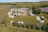An aerial view of a campsite filled with numerous RVs and caravans. The area is surrounded by lush green grass and trees, with a river seen in the background. Vehicles are parked in an organized manner around a central dirt path, and there are people visible near their camping setups. The landscape is open and expansive, with a few scattered houses and hills in the distance.