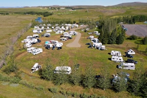 An aerial view of a campsite filled with numerous RVs and caravans. The area is surrounded by lush green grass and trees, with a river seen in the background. Vehicles are parked in an organized manner around a central dirt path, and there are people visible near their camping setups. The landscape is open and expansive, with a few scattered houses and hills in the distance.