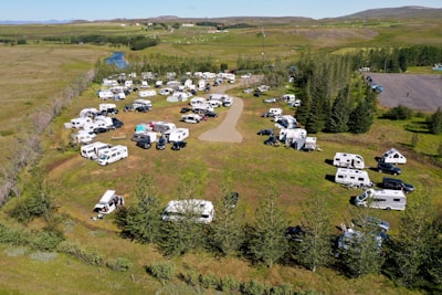 An aerial view of a campsite filled with numerous RVs and caravans. The area is surrounded by lush green grass and trees, with a river seen in the background. Vehicles are parked in an organized manner around a central dirt path, and there are people visible near their camping setups. The landscape is open and expansive, with a few scattered houses and hills in the distance.