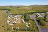 Aerial view of an expansive campground with numerous recreational vehicles and campers parked across grassy fields. A winding river runs in the background alongside open green landscapes. Several trees are scattered across the area, and buildings can be seen on the right side.