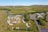 Aerial view of an expansive campground with numerous recreational vehicles and campers parked across grassy fields. A winding river runs in the background alongside open green landscapes. Several trees are scattered across the area, and buildings can be seen on the right side.