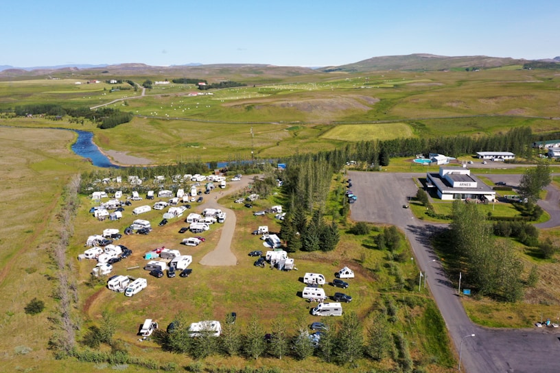 Aerial view of an expansive campground with numerous recreational vehicles and campers parked across grassy fields. A winding river runs in the background alongside open green landscapes. Several trees are scattered across the area, and buildings can be seen on the right side.