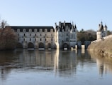 A large, elegant chateau spans a river, showcasing arches that reflect on the calm water. The architecture features towers and intricate details, surrounded by lush greenery and trees. The sky is clear and blue.