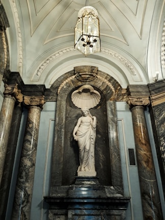 A white marble statue of a classical figure stands in an ornate niche with a shell-shaped backdrop. The statue is surrounded by grand marble columns with intricate capitals, and the ceiling is elaborately decorated with arches and a chandelier.
