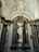 A white marble statue of a classical figure stands in an ornate niche with a shell-shaped backdrop. The statue is surrounded by grand marble columns with intricate capitals, and the ceiling is elaborately decorated with arches and a chandelier.