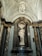 A white marble statue of a classical figure stands in an ornate niche with a shell-shaped backdrop. The statue is surrounded by grand marble columns with intricate capitals, and the ceiling is elaborately decorated with arches and a chandelier.