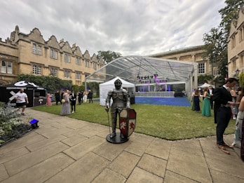 A historical building with ornate architecture serves as the backdrop for an outdoor event. People dressed in formal attire are gathered in a courtyard. A knight's armor stand with a shield is positioned prominently in the foreground. A transparent tent with purple and blue lighting is set up for an event, and a small stand labeled 'Grosstown Doughnuts' can be seen on the left.