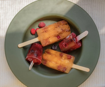 Three colorful popsicles with visible pieces of fruit are arranged on a round green plate. The popsicles are made of red and orange translucent ice, containing small berries. A few loose berries surround the popsicles, adding to the vibrant presentation. Sunlight casts soft shadows, enhancing the colorful appeal.