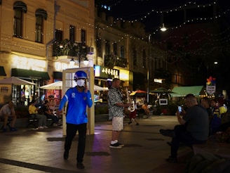 A lively street scene at night with people sitting and interacting. A person in a blue outfit and helmet walks in the foreground. Another individual plays a saxophone, surrounded by an audience, while someone captures the moment on a smartphone. Buildings with illuminated signs and string lights create a vibrant atmosphere.