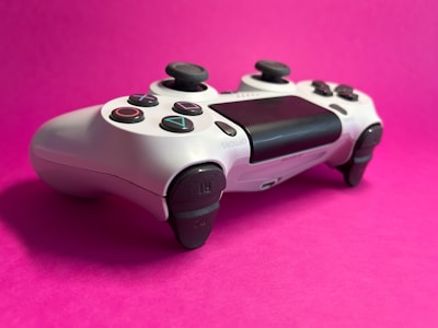 A white game controller with black buttons and joysticks placed on a vibrant pink background. The symbols on the buttons are clearly visible, and the device is positioned to highlight its ergonomic design and features.