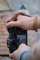 A close-up of a person's hands holding a camera with a brown strap wrapped around the wrist. The camera's display screen shows a live view of a house with a pointed roof and chimney. The background is out of focus, showing a tiled roof surface.