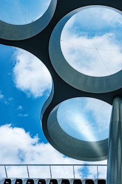Circular glass panels embedded in a metallic framework create an abstract architectural design against a backdrop of a blue sky with scattered clouds. The scene is dominated by geometric shapes and a futuristic aesthetic.