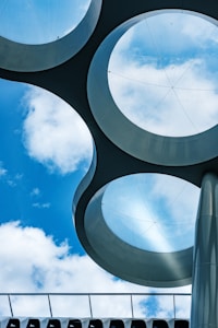 Circular glass panels embedded in a metallic framework create an abstract architectural design against a backdrop of a blue sky with scattered clouds. The scene is dominated by geometric shapes and a futuristic aesthetic.