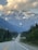 A scenic highway stretches into the distance with two semi-trucks driving toward snow-capped mountains partly covered by clouds. Dense forest lines both sides of the road.
