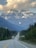 A scenic highway stretches into the distance with two semi-trucks driving toward snow-capped mountains partly covered by clouds. Dense forest lines both sides of the road.