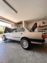 A classic sedan is parked inside a garage, raised on a jack with the rear wheel removed. The vehicle's trunk and hood are open, while shelves filled with various tools and car parts line the background. A partially visible person appears to be working on the car.