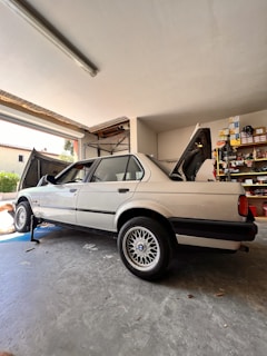A classic sedan is parked inside a garage, raised on a jack with the rear wheel removed. The vehicle's trunk and hood are open, while shelves filled with various tools and car parts line the background. A partially visible person appears to be working on the car.