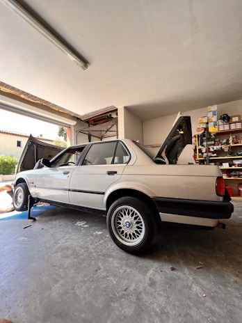 A classic sedan is parked inside a garage, raised on a jack with the rear wheel removed. The vehicle's trunk and hood are open, while shelves filled with various tools and car parts line the background. A partially visible person appears to be working on the car.