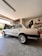 A classic sedan is parked inside a garage, raised on a jack with the rear wheel removed. The vehicle's trunk and hood are open, while shelves filled with various tools and car parts line the background. A partially visible person appears to be working on the car.