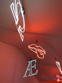 Neon lights hang from the ceiling, showcasing different shapes and letters. A hand outline is formed in bright orange neon, while letters such as 'AE' are depicted in white and blue neon. The lights are suspended against a warm orange background.