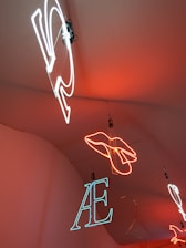 Neon lights hang from the ceiling, showcasing different shapes and letters. A hand outline is formed in bright orange neon, while letters such as 'AE' are depicted in white and blue neon. The lights are suspended against a warm orange background.