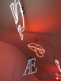 Neon lights hang from the ceiling, showcasing different shapes and letters. A hand outline is formed in bright orange neon, while letters such as 'AE' are depicted in white and blue neon. The lights are suspended against a warm orange background.