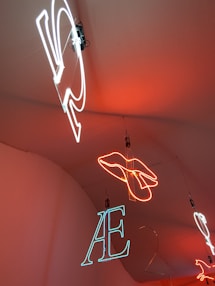 Neon lights hang from the ceiling, showcasing different shapes and letters. A hand outline is formed in bright orange neon, while letters such as 'AE' are depicted in white and blue neon. The lights are suspended against a warm orange background.