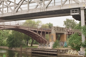 A two-level bridge with steel structures crosses over a body of water adorned with graffiti. The word 'BERLIN' is prominently displayed on the lower part of the bridge. Surrounding the bridge are trees and a small waterway, contributing to a blend of urban and natural elements.