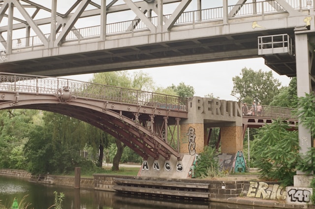 A two-level bridge with steel structures crosses over a body of water adorned with graffiti. The word 'BERLIN' is prominently displayed on the lower part of the bridge. Surrounding the bridge are trees and a small waterway, contributing to a blend of urban and natural elements.