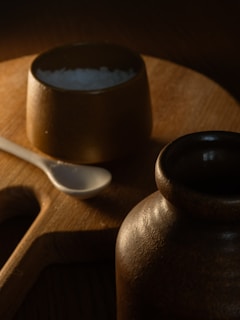 A wooden cutting board holds a small brown ceramic bowl filled with coarse salt and a white ceramic spoon placed beside it. In the foreground, a larger brown ceramic jar is partially visible, with a soft, warm lighting creating a shadowy and rustic atmosphere.