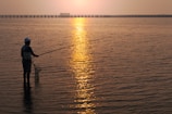A young adult wearing a wide-brimmed hat, casting a fishing line into a calm Florida bay at sunset.
