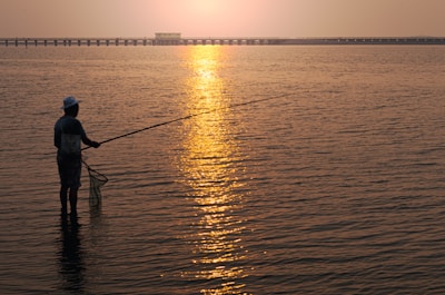 A young adult wearing a wide-brimmed hat, casting a fishing line into a calm Florida bay at sunset.