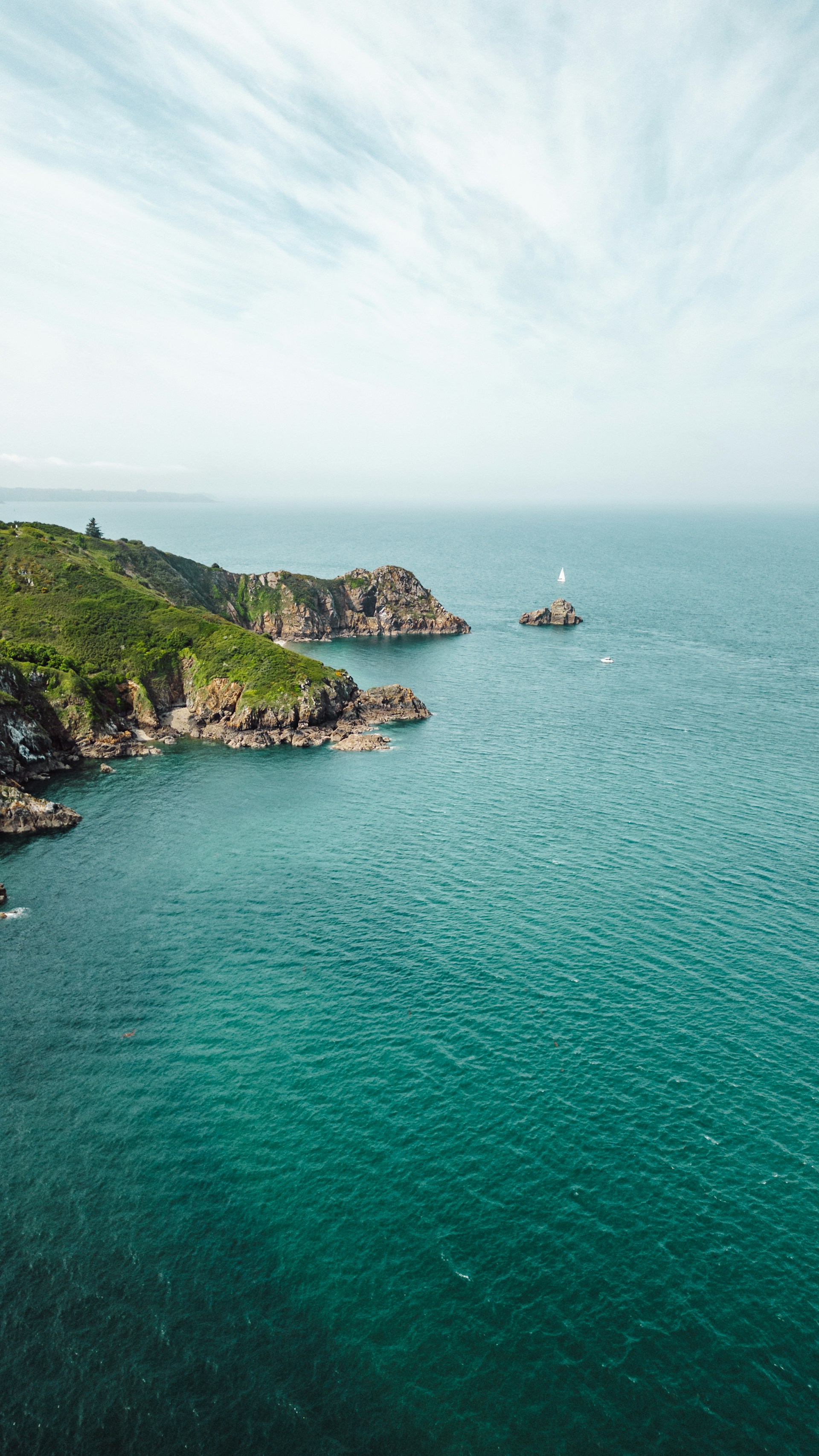 A sunlit coastal cliff overlooking turquoise waves with a small sailboat drifting nearby.