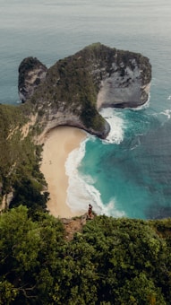 A stunning coastal landscape featuring a horseshoe-shaped cliff covered in lush greenery. The turquoise ocean waves gently lap against the secluded sandy beach below. A lone person stands on the edge of the cliff, taking in the panoramic view.