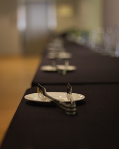 A table is set with white plates and utensils, arranged neatly on a black tablecloth. The setting creates a sense of elegance and formality, with a shallow depth of field causing the background to be out of focus.